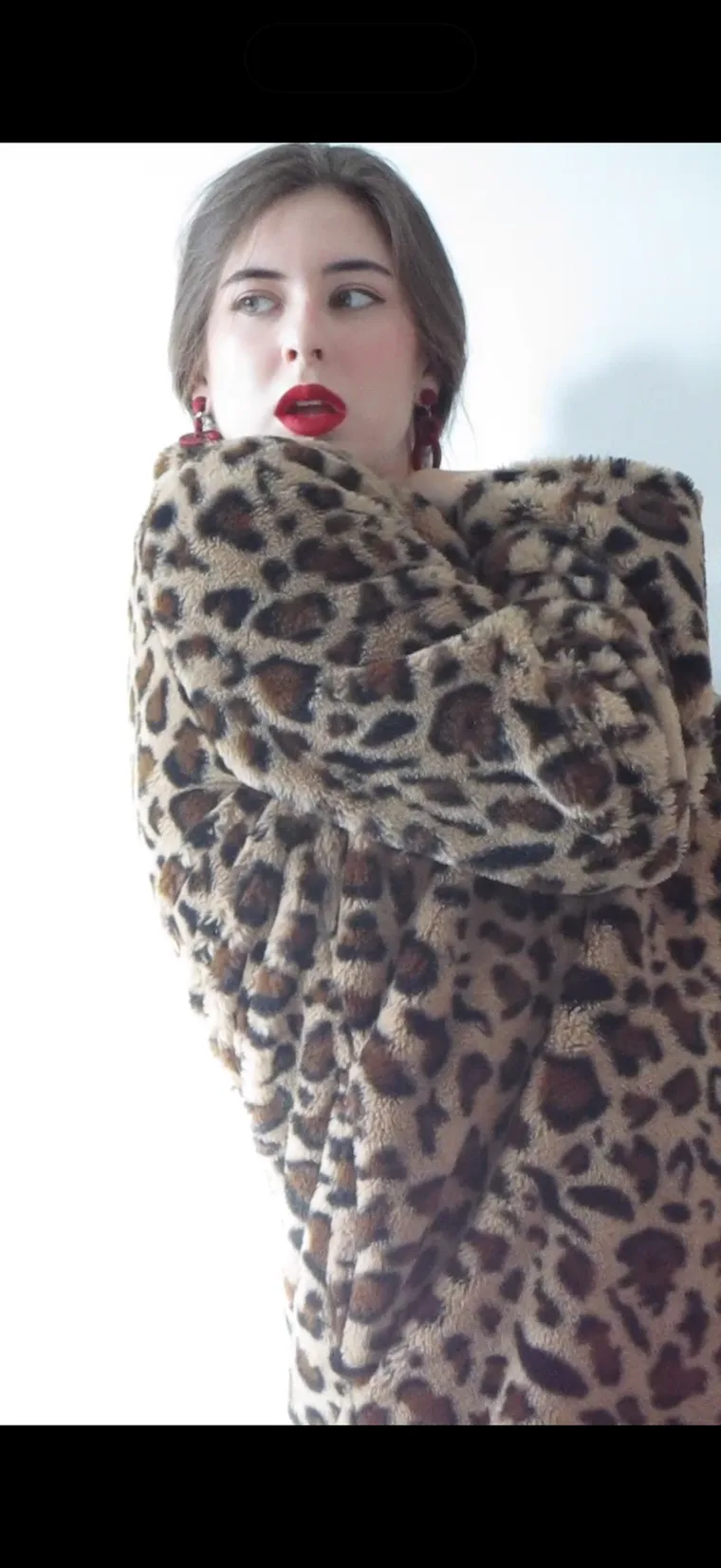 Virginia Kate Brandt in an oversized leopard-print coat, looking off-camera, wearing red earrings and red lipstick.