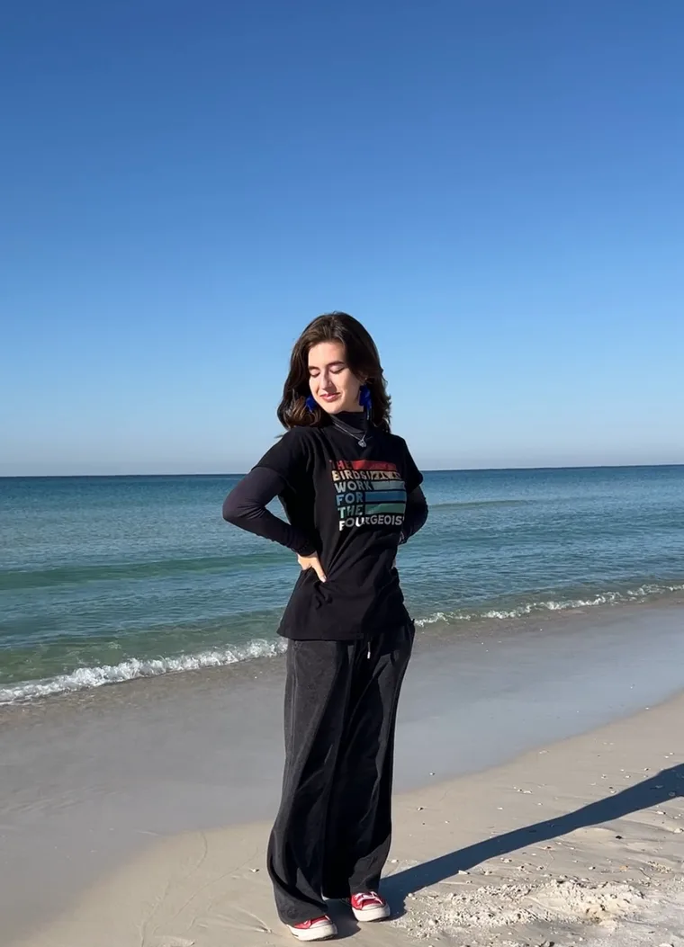 Portrait on the beach in a black 'Birds Work For The Bourgeoisie' long-sleeve, blue tassel earrings, wide-leg pants and red sneakers.