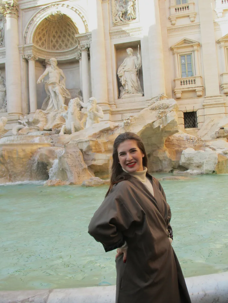 Portrait in a chocolate-brown coat over an ivory turtleneck, in front of the Trevi Fountain, Rome.