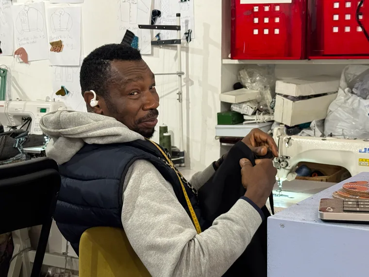 Earnest Alulu at his sewing machine in the Coloriage Laboratorio workroom, holding a dark garment under the needle.